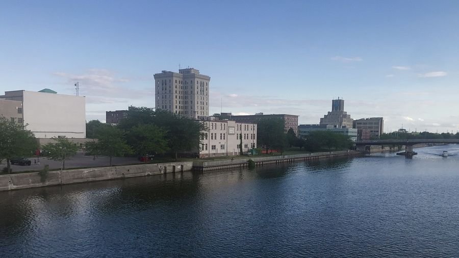 The Saginaw River on a sunny day, with trees along the riverbank and historic downtown Saginaw buildings in the background.